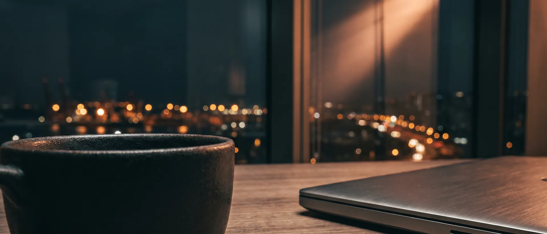 Borde de una laptop cerrada y una taza frente a una ventana con la ciudad de noche